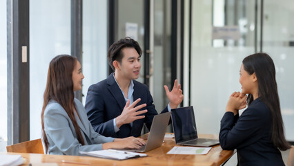 Three business professionals engaged in a meeting around a table, discussing projects with laptops in an office setting.
