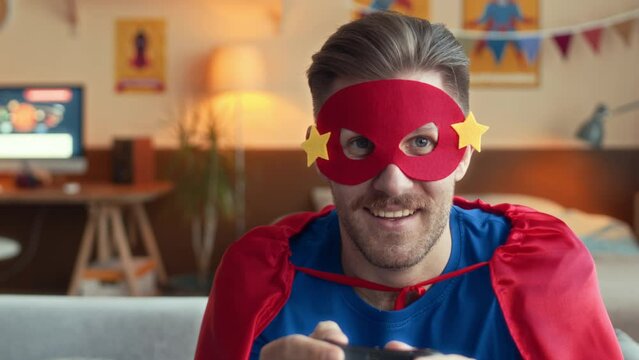 Medium shot of excited Caucasian young adult man wearing red superhero eye mask and costume having fun playing video games at home with son