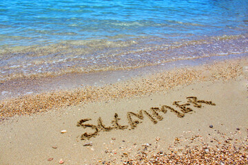 The word summer is written in the sand next to a beautiful blue ocean with waves lapping on the shore