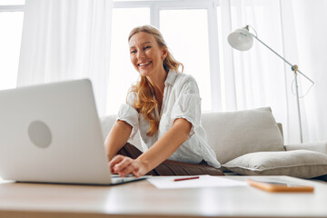 Smiling woman sitting on a couch with a laptop, working comfortably from home in a cozy living room setting