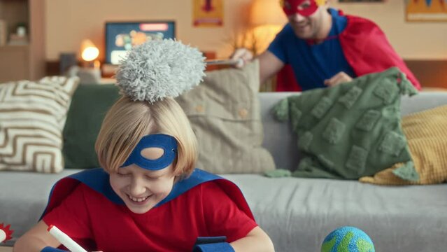 Playful Caucasian man stroking tween sons head with pp duster while having fun at home together both dressed in blue and red superhero costumes