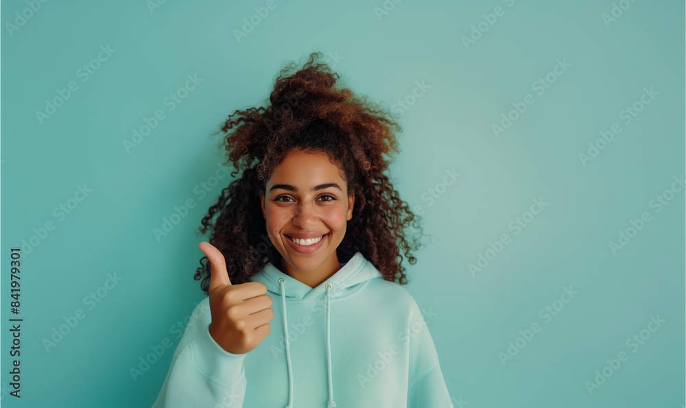 Wall mural Young curly african american girl in a sweatshirt smiling with healthy teeth showing thumb up at copy space expressing wow emotion standing isolated on mint background - Wall murals