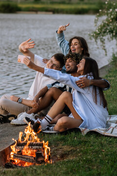 Company of young friends having fun and taking selfies in nature on the background of a lake sitting by a fire.