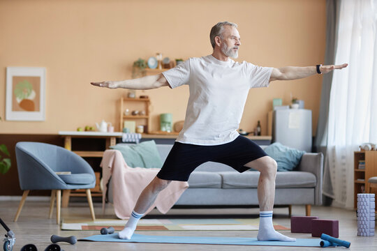 Full length side view of strong senior man standing in warrior pose during yoga practice at home copy space