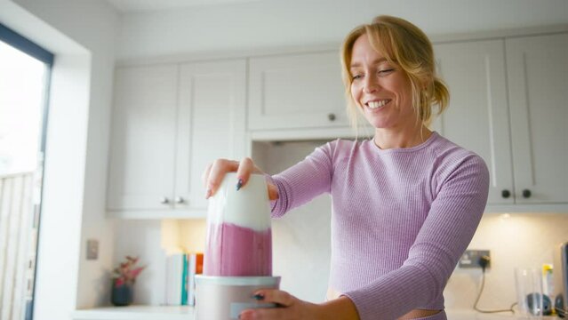 Woman wearing fitness clothing at home in kitchen making healthy smoothie mixing up yoghurt, banana,raspberries and blueberries in blender- shot in slow motion
