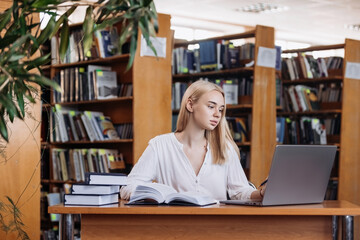 Serious hardworking blonde female student watching webinar, listening online learning course, looking at laptop display, writing notes at table in college library