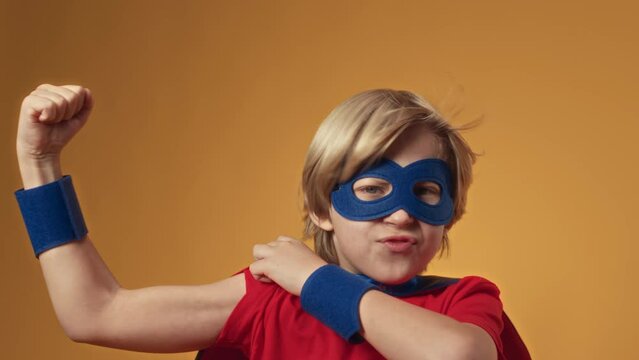 Medium portrait of Caucasian 9 year old boy wearing red and blue superhero costume showing muscles at camera and wind blowing his hair and cloak