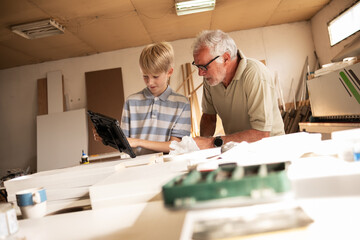 Obraz premium Grandson teaching his grandfather carpenter how to use a tablet to improve their work. The grandson shares his tech knowledge, helping the grandfather integrate modern tools into their craft.