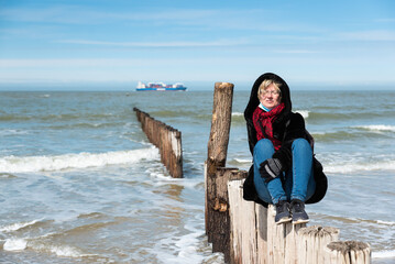 30 yo white woman with red scarf, winter jacket sitting on a breakwater pilar in the sun at the beach, The Netherlands