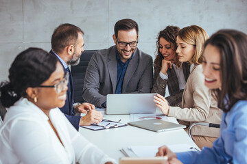 A group of professional men and women of different ethnicities actively participate in a business meeting, discussing over a laptop in a modern office setting.