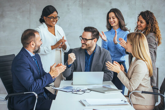 A group of diverse business professionals celebrate success, clapping for a smiling male colleague seated with a laptop in a modern office.