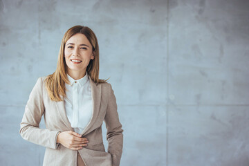 A professional Caucasian woman smiles engagingly, dressed in a chic blazer, poised confidently in a well-lit office with a minimalist industrial wall.