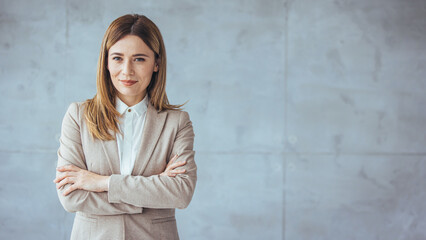 A professional Caucasian woman stands with arms crossed wearing business attire in an office setting, featuring a stylish industrial wall backdrop.