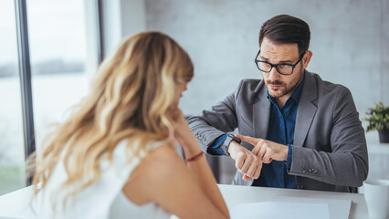 A focused Caucasian businesswoman and businessman review documents and discuss strategies while sitting at a desk in a well-lit office, utilizing time management.
