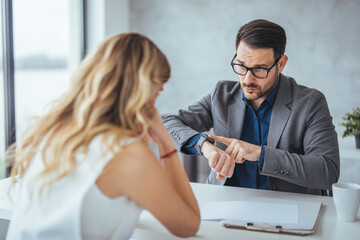 A focused Caucasian businesswoman and businessman engage in collaboration at an office desk, discussing work with documents and a wristwatch in view.