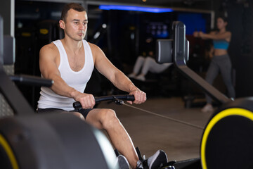 Sportive young man doing exercises on cable rowing device in well-equipped gym