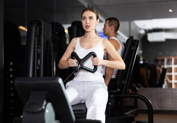 Sportive young woman doing arm exercises on cable training device in well-equipped gym