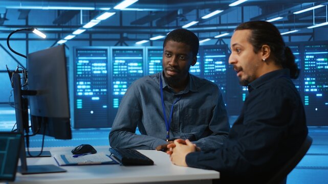 Multiracial engineers programming in high tech facility with server rows providing computing resources for different workloads. Diverse coworkers supervising data center supercomputers
