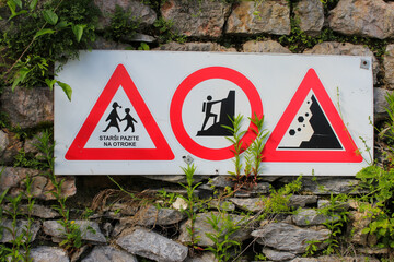 There is a sign with three warning road signs hanging on a stone wall overgrown with green grass.