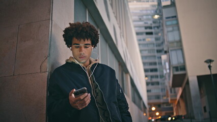Latino guy scrolling mobile phone on city street wearing earphones closeup. 