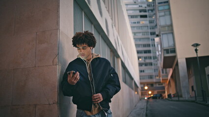 Urban guy checking smartphone standing street at dusk in headphones. Latino man