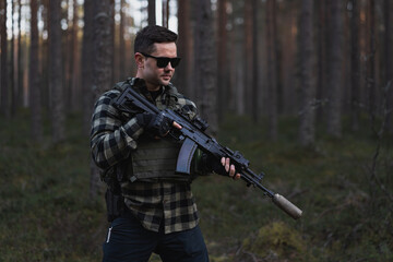A male mercenary in a bulletproof vest over his shirt, wearing sunglasses and holding an assault...