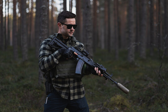 A male mercenary in a bulletproof vest over his shirt, wearing sunglasses and holding an assault rifle in his hands in the forest.