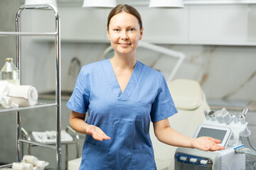 Portrait of middle-aged smiling masseuse doctor woman standing near beauty machine in modern medical salon, stretching hands aside with open palm, inviting clients
