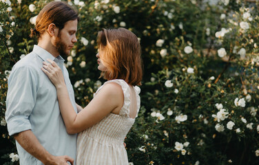 Fototapeta premium beautiful couple in retro clothes hugging near a tree of white roses in the summer in the village.