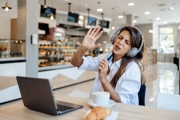 Beautiful and happy young woman sitting and eating delicious rolls in bakery or fast food. She also using her laptop and listening to music with headphones.