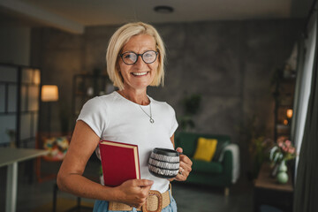 Portrait of senior mature woman stand and hold cup and book at home