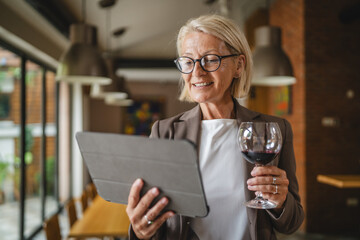Senior woman stand hold glass of wine and use tablet in the winery