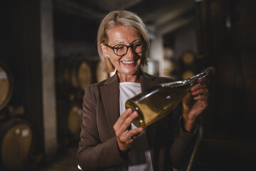Mature senior woman winemaker stand in basement hold bottle of wine