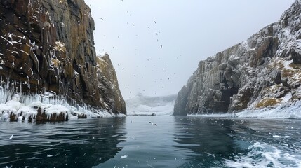 Birds soar over icy waters surrounded by imposing cliffs in a remote Arctic seascape with overcast...
