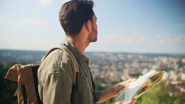 Zooming out from handsome man with backpack fore travel looking at map while standing on top of cliff. Young hiker exploring new place. Observing beautiful view of city. Relaxation on wild nature.