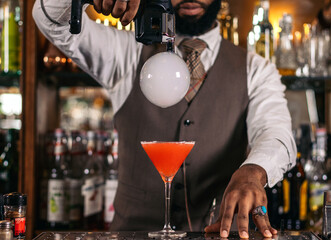 Stylish black bartender preparing a cocktail with a smoke bubble in a traditional cocktail bar