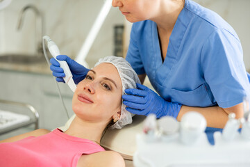 Caucasian brunette young girl lying in spa while having facial treatment procedures with modern...