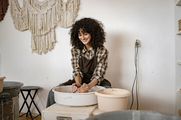 Female potter using wheel to sculpt ceramic bowl in cozy studio. Curly haired woman enjoying...