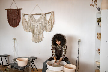 Female artist working on ceramic dish using potter's wheel in a decorative studio with wall...