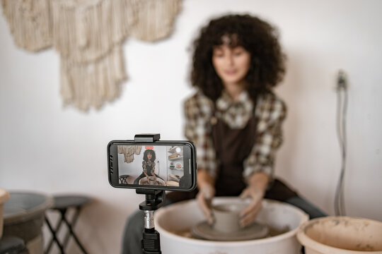 Young female potter sculpting a jug using a potter's wheel while recording a video blog with a mobile phone. Creative art process and vlogging.
