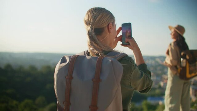 Camera moving around two Caucasian women. People standing on top of hill while taking photos on cell prone of each other. Using smartphone to record images. Concept of active lifestyle.