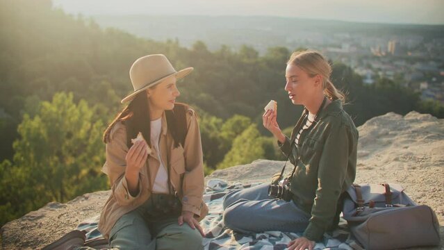 Two Caucasian women sitting on blanket on edge of cliff. Enjoying their meal as sun shines bright during sundown. Females laughing and smirking to each other. Camping after hard hike.