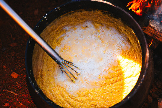 A large whisk in a cast iron pot of hot yellow grits cooking over an open fire.