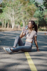 A young woman in athletic wear sitting on a park path, drinking water, and enjoying a sunny day.