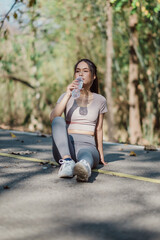 A young woman in sportswear sits on a pathway in a forest, drinking water and taking a break from...