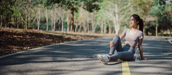 Young woman in athletic wear sitting on a forest road, resting after a workout, with trees and...