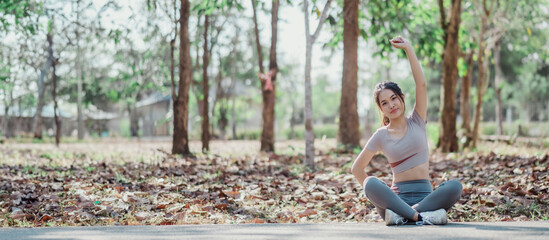 A woman is doing yoga in a park. She is wearing a grey shirt and grey pants. The park is filled with trees and the ground is covered in leaves. The woman is in a relaxed