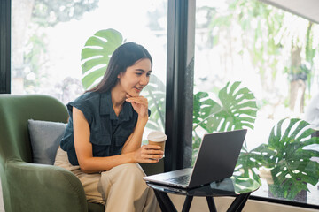 A woman is sitting in a chair with a laptop open in front of her. She is holding a cup of coffee and smiling. Concept of relaxation and productivity