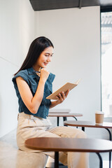 A woman is sitting in a cafe reading a book. She is smiling and she is enjoying her time. The cafe has wooden tables and chairs, and there are cups and a bottle on the tables