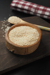A wooden bowl filled with white sesame seeds, accompanied by a spoon, set on a dark surface